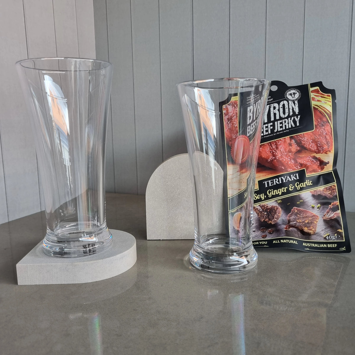 Two unbreakable DStill Pilsner Beer Glasses, Aureliia Collection Arch Coasters in Airy Concrete with a Byron Beef Jerky package in the background.