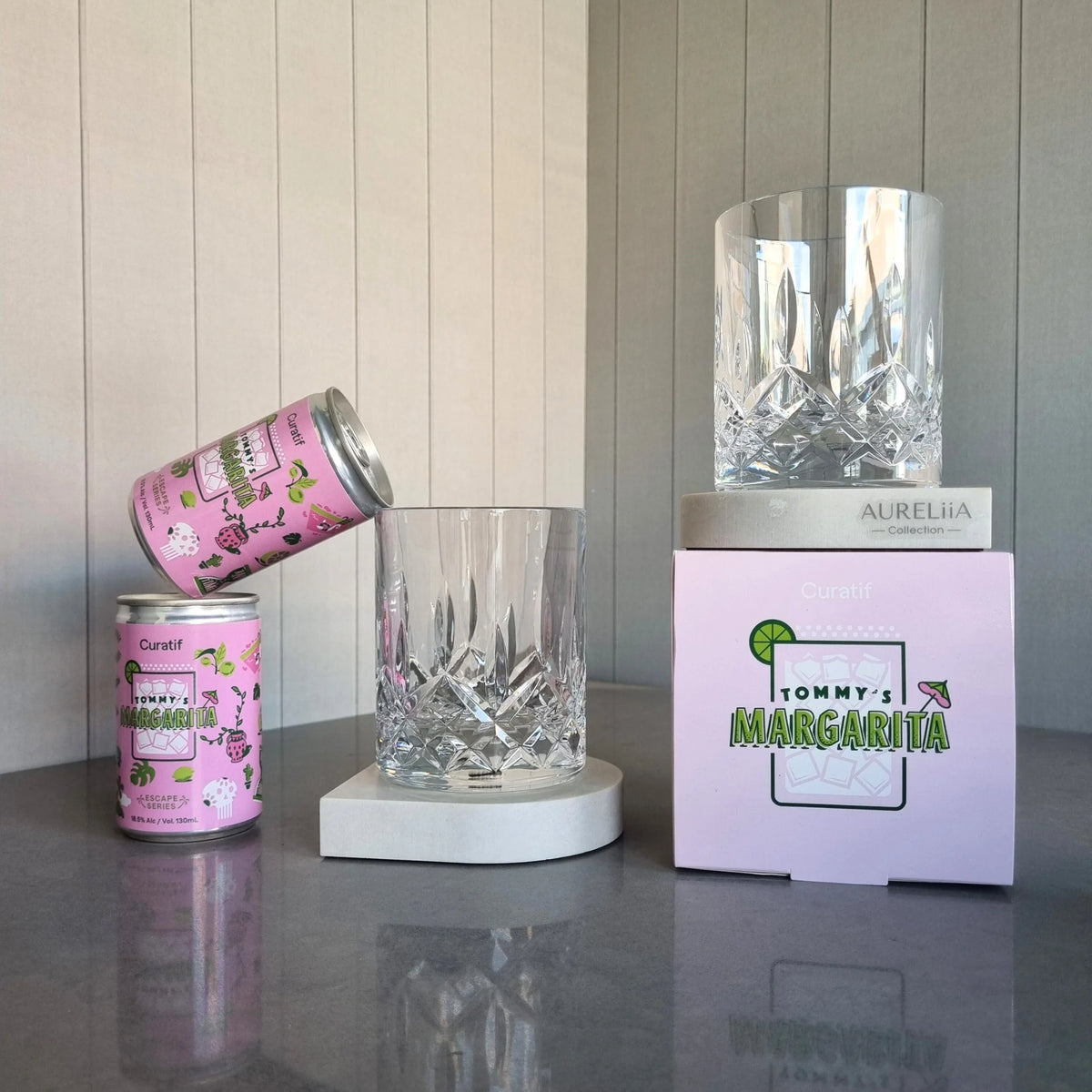 Two DStill unbreakable diamond cut glass tumblers on marble coaster arch shape in Caesarstone Alpine Mist and a pink canister Curatif Tommy's Margarita on a reflective surface with a neutral background. Gifts for Dad.