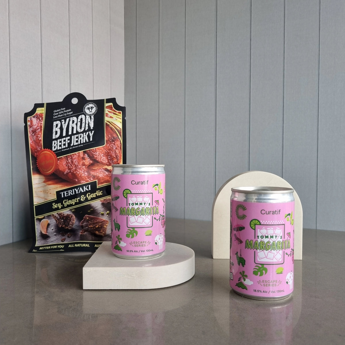 Two cans of Tommy's Margarita on a gray surface with a package of Byron Beef Jerky in the background.