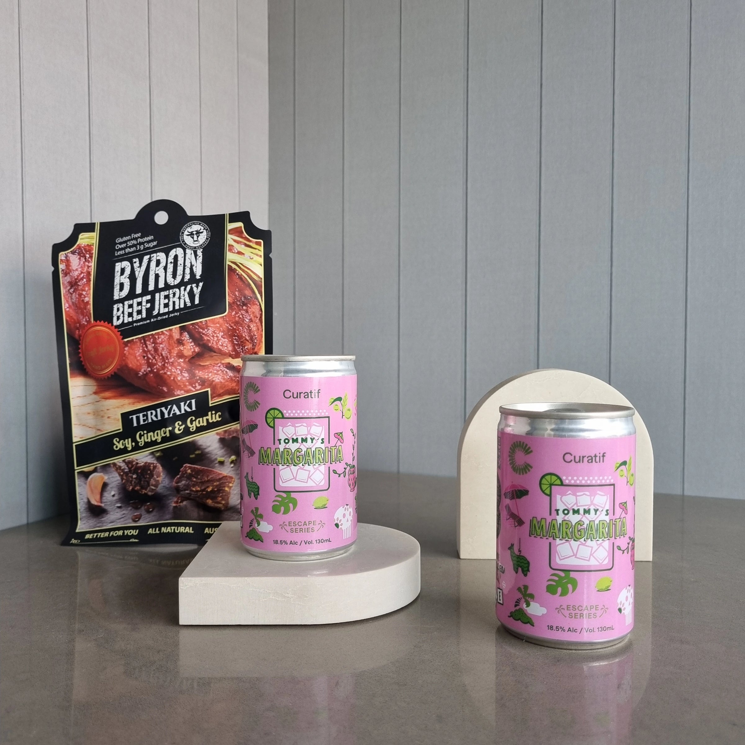 Two cans of Tommy's Margarita on a gray surface with a package of Byron Beef Jerky in the background.