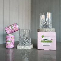 Two cans of Curatif Tommy's Margarita and two DStill unbreakable crystal glasses with arch coasters on a grey reflective surface with a striped wall background.