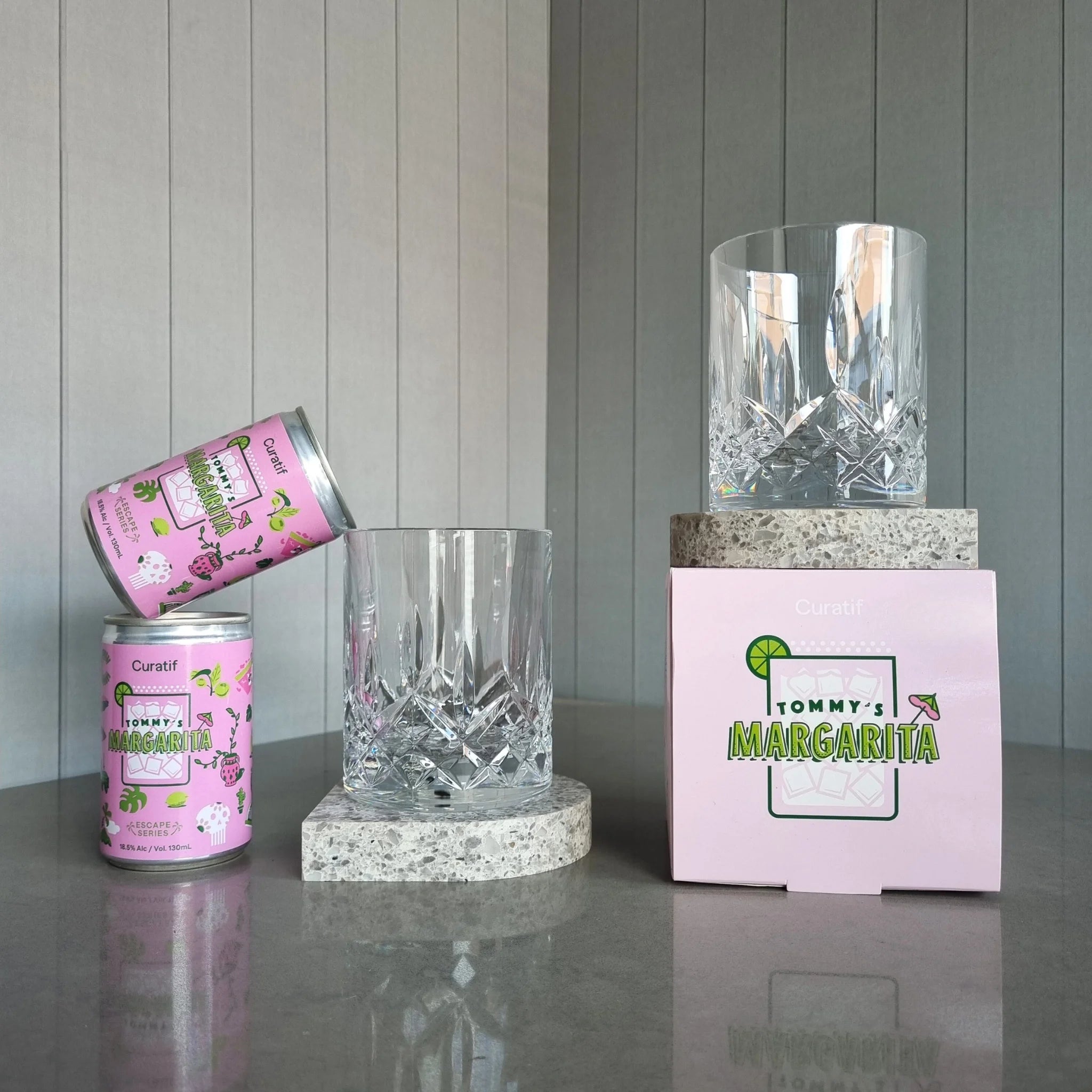 Two cans of Curatif Tommy's Margarita and two DStill unbreakable crystal glasses with arch coasters on a grey reflective surface with a striped wall background.