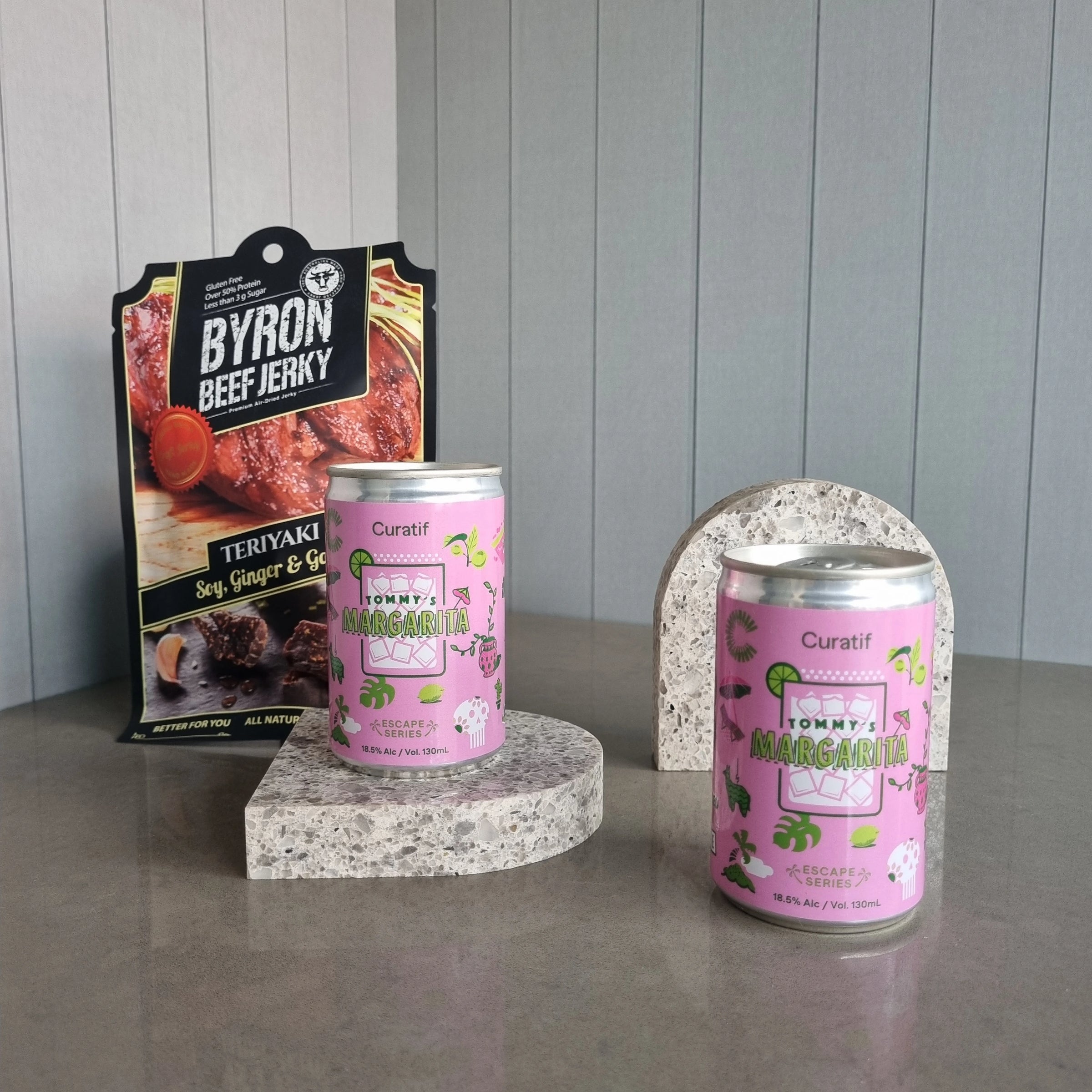 Two cans of 'Curatif' margarita on stone stands with a package of 'Byron Beef Jerky' in the background.