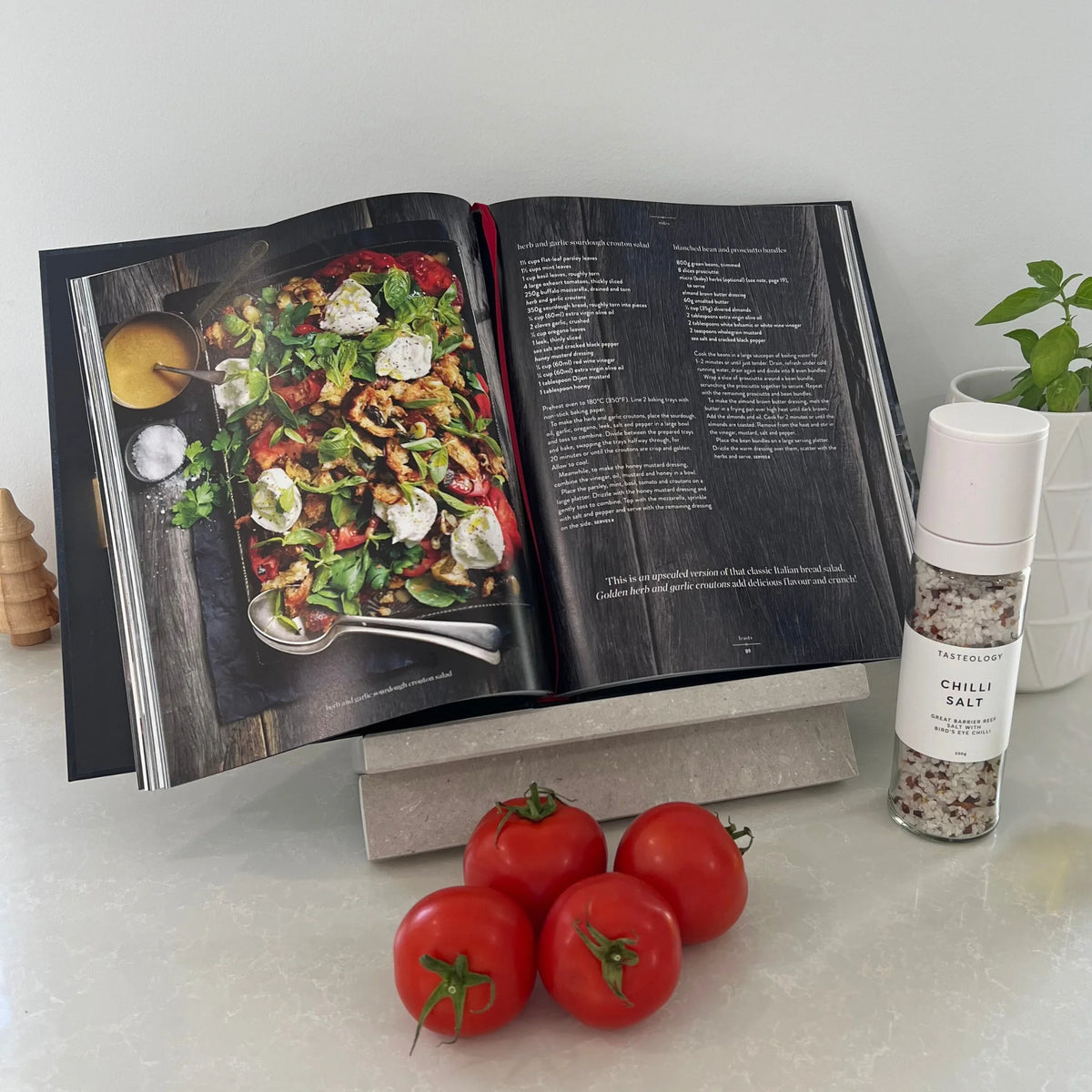 Open cookbook with a dish on a marble cookbook stand, surrounded by tomatoes and a Tasteology salt.