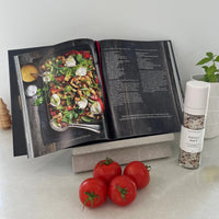 Open cookbook with a dish on a marble cookbook stand, surrounded by tomatoes and a Tasteology salt.