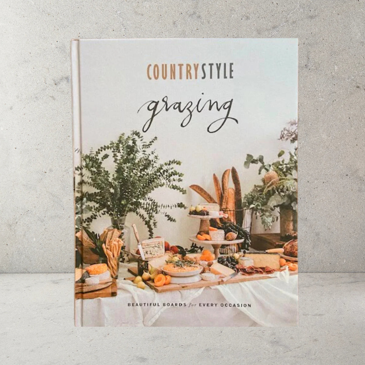 The Australian Womens Weekly Country Style Grazing Cookbook with hard cover. Shows table set with grazing board, native greenery and fresh bread.