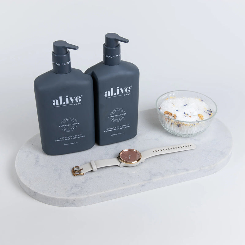 Marble double arch tray holding two black pump bottles of alive brand liquid hand soap and hand cream, and a glass bowl with a bath salts. In front of these items is a garmin watch with a gold band.