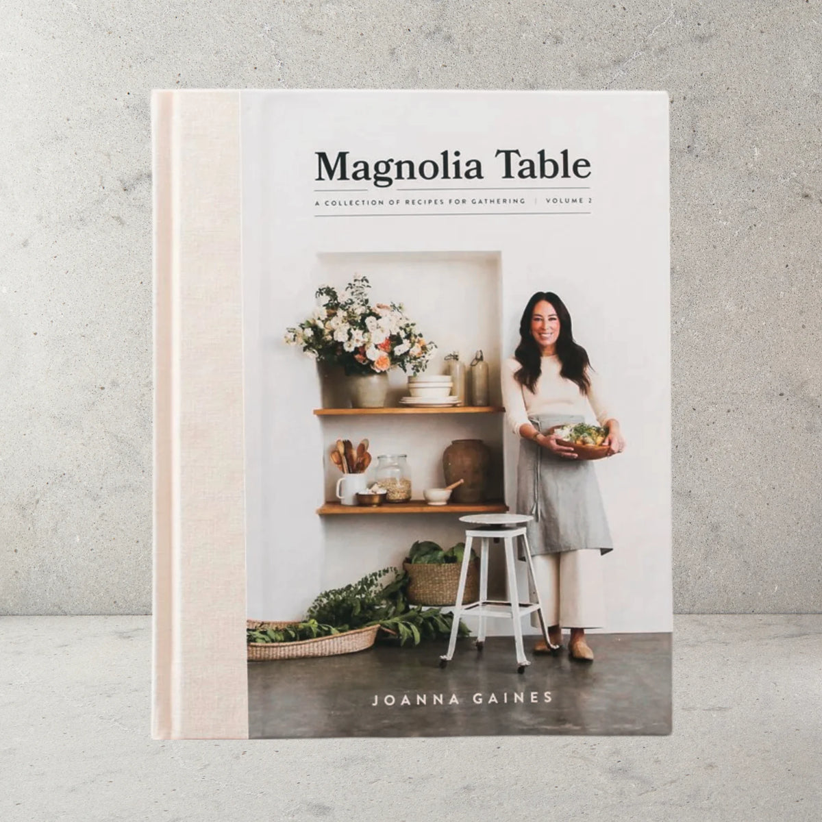 Magnolia Table Cookbook with hard cover by Joanna Gaines. Cover show picture of Joanna with fresh flowers, styled shelves, utensils and more.