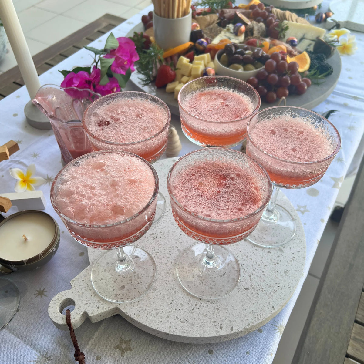 Pink cocktails in glasses on Christmas shaped platter with a charcuterie board and candles.