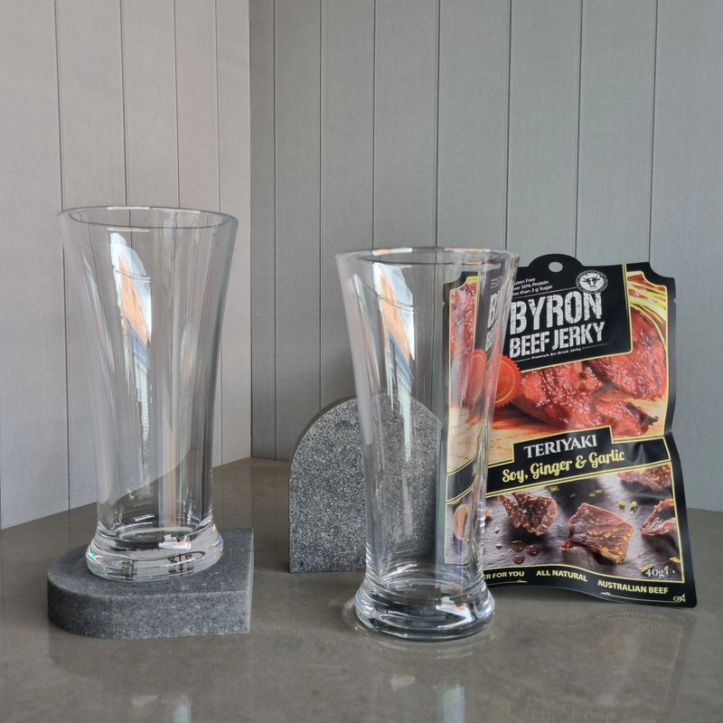 Two DStill Pilsner Beer Glasses on a grey surface, Aureliia Collection Arch Coasters in Caesarstone Rugged Concrete with a Byron Beef Jerky package in the background.