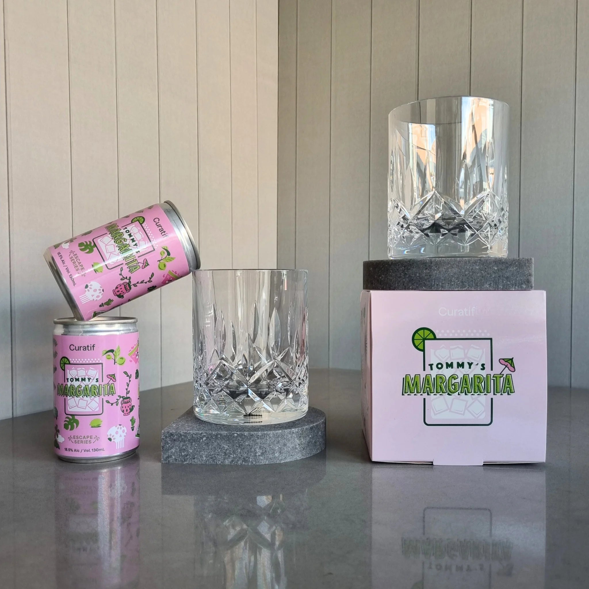 Two cans of Curatif Tommy's Margarita and two DStill unbreakable glasses on a grey Caesarstone Rugged Concrete stone coasters on reflective surface with a striped wall background.