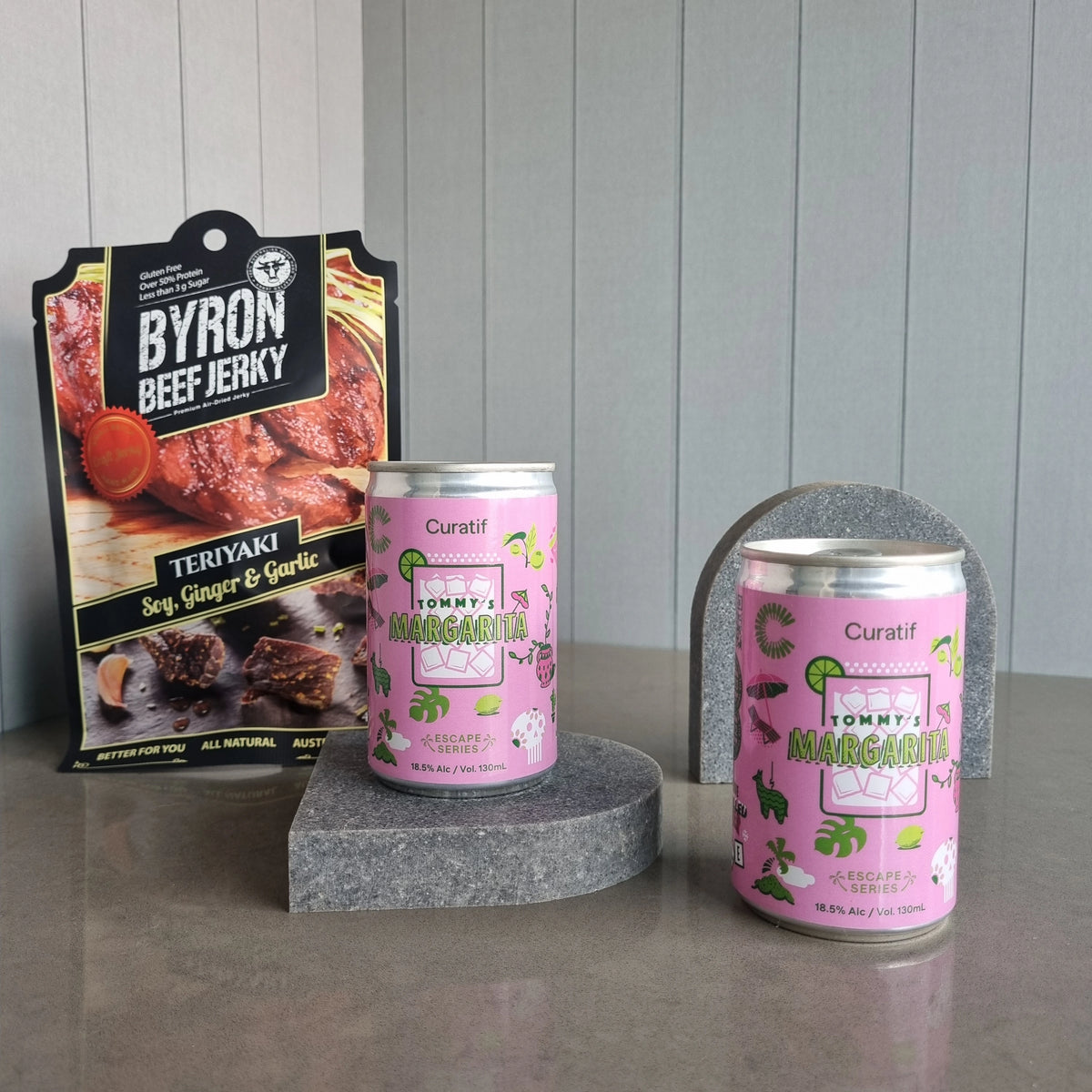 Two cans of Tommy's Margarita on a gray surface with a package of Byron Beef Jerky in the background.