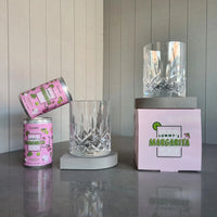 Two cans of Curatif Tommy's Margarita and two DStill unbreakable glasses on a grey Caesarstone arch coasters on reflective surface with a striped wall background.