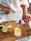 Person pouring a can of Curatif Tommy's Margarita into a glass with lime slices cut on timber chopping board with wicker placemat. Father’s Day gifts.