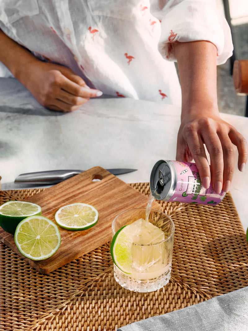 Person pouring a can of Curatif Tommy's Margarita into a glass with lime slices cut on timber chopping board with wicker placemat. Father’s Day gifts.